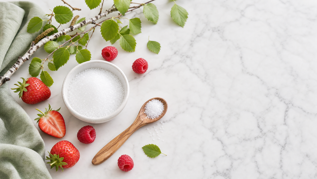 A bowl of xylitol crystals with birch branches, strawberries, and raspberries on a marble surface