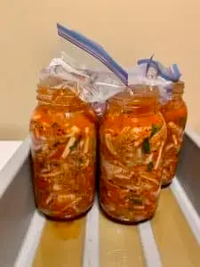 A two quart jar of kimchi fermenting on the counter with a water filled bag holding the cabbage under the brine