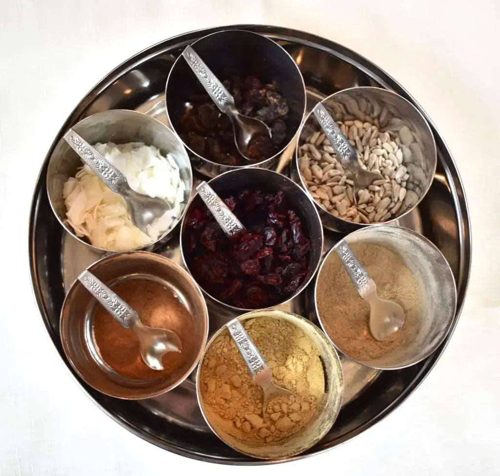 A stainless steel tiffin box filled with small cups of raisins, dried cranberries, sunflower seeds, coconut and warming spices for topping hot cereal