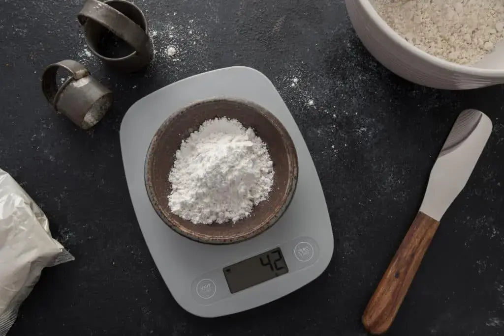 Flour weighed in grams on a digital kitchen scale next to a bowl of flour and a dough scraper